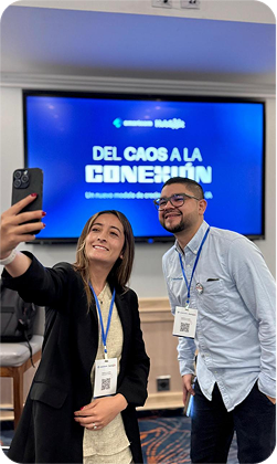 Dos personas del equipo se toman una selfie en un evento de Smarteam, frente a una pantalla que dice «Del caos a la conexión».